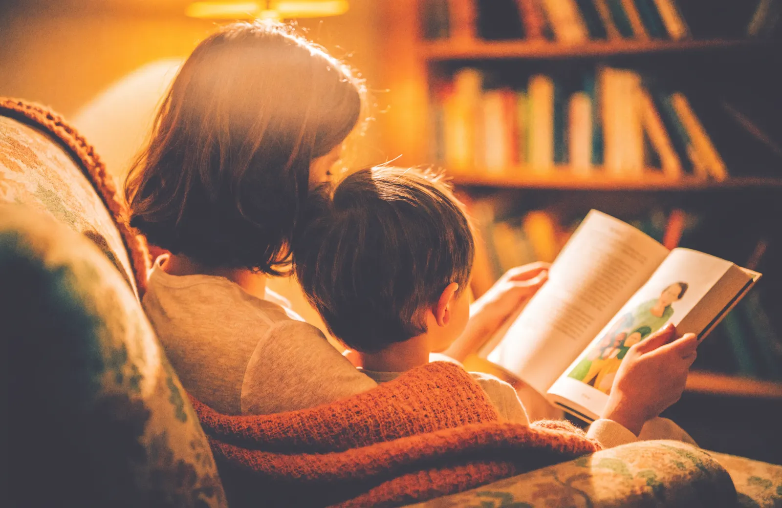 A parent and child seen from behind, reading a picture book together in an armchair by lamplight.